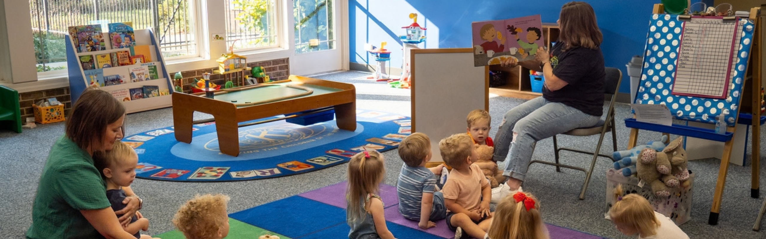 Two women with several toddlers and young children in a brightly lit room with a story time and play area setup.